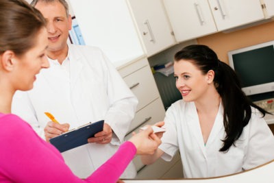 Patient in reception area of office of doctor or dentist, handing her health insurance card over the counter to the nurse, the doctor standing in the background and is writing things on a clipboard