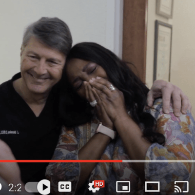 Mature black woman puts her hands to her face in a gesture of shock and is hugged by a dentist